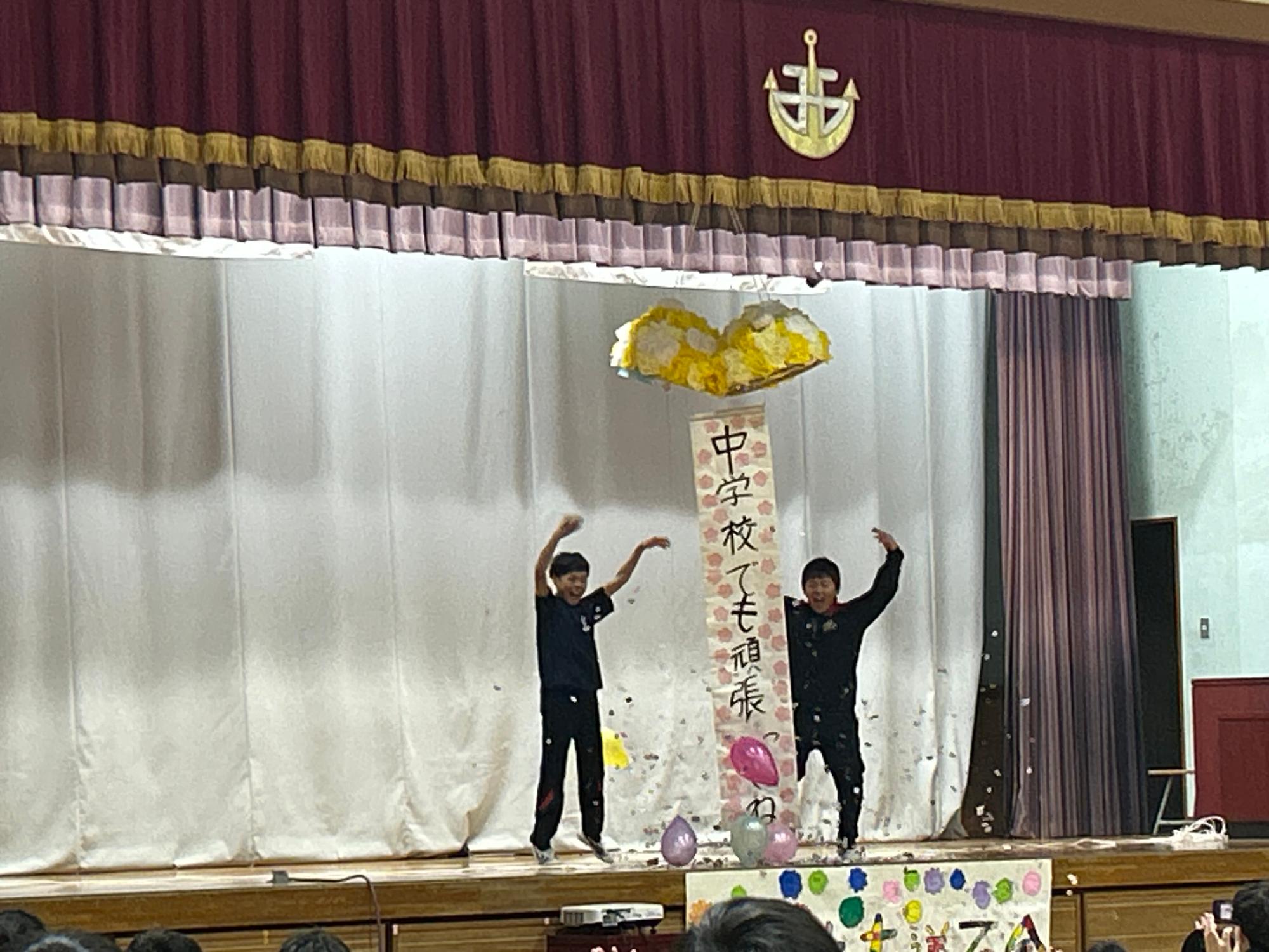 体育館の舞台上で児童たちがくす玉を割ってメッセージを発表している写真