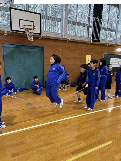 体育館で子どもたちが大縄跳びをしており、それを複数の児童が座ってみている様子の写真