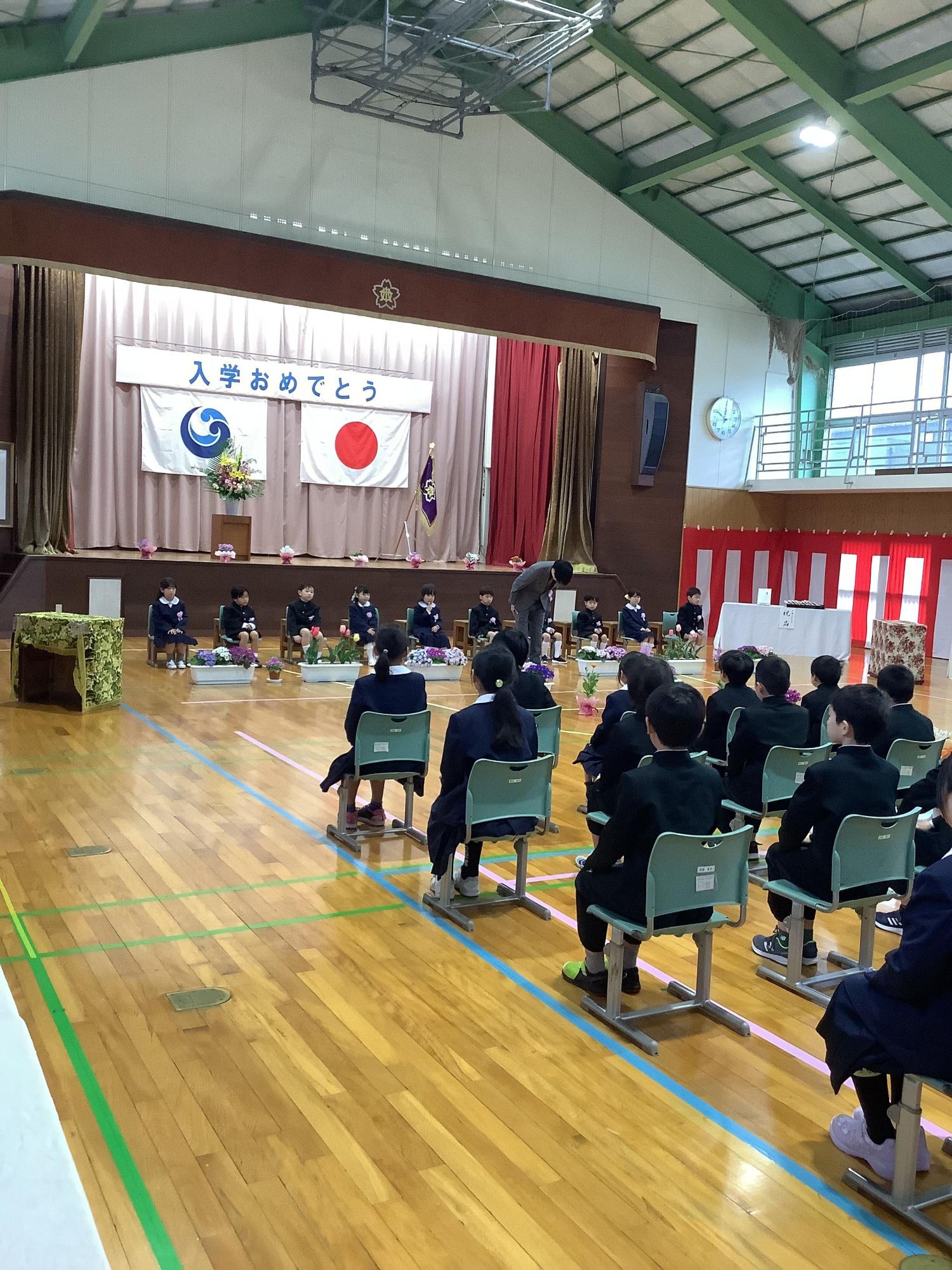 体育館の舞台前で新入生の児童たちが入学式に臨んでいる写真