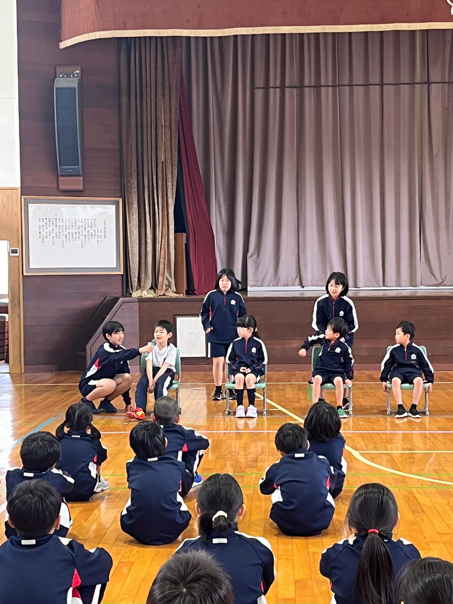 体育館で児童たちが椅子に座り、上級生からインタビューを受けている写真