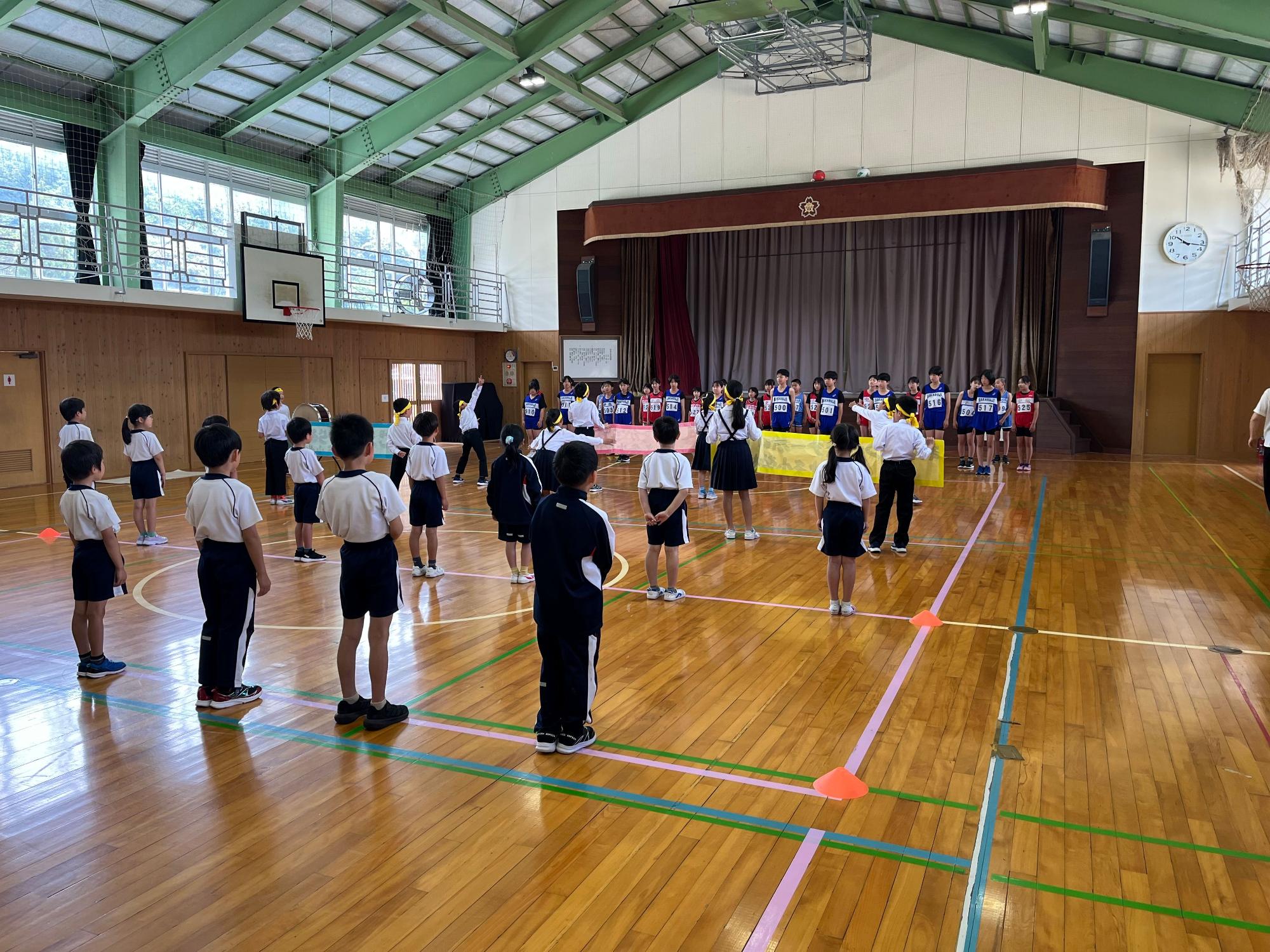 体育館でジャージ姿の児童たちとマラソンのユニフォームを着た児童たちが向かい合って整列している写真