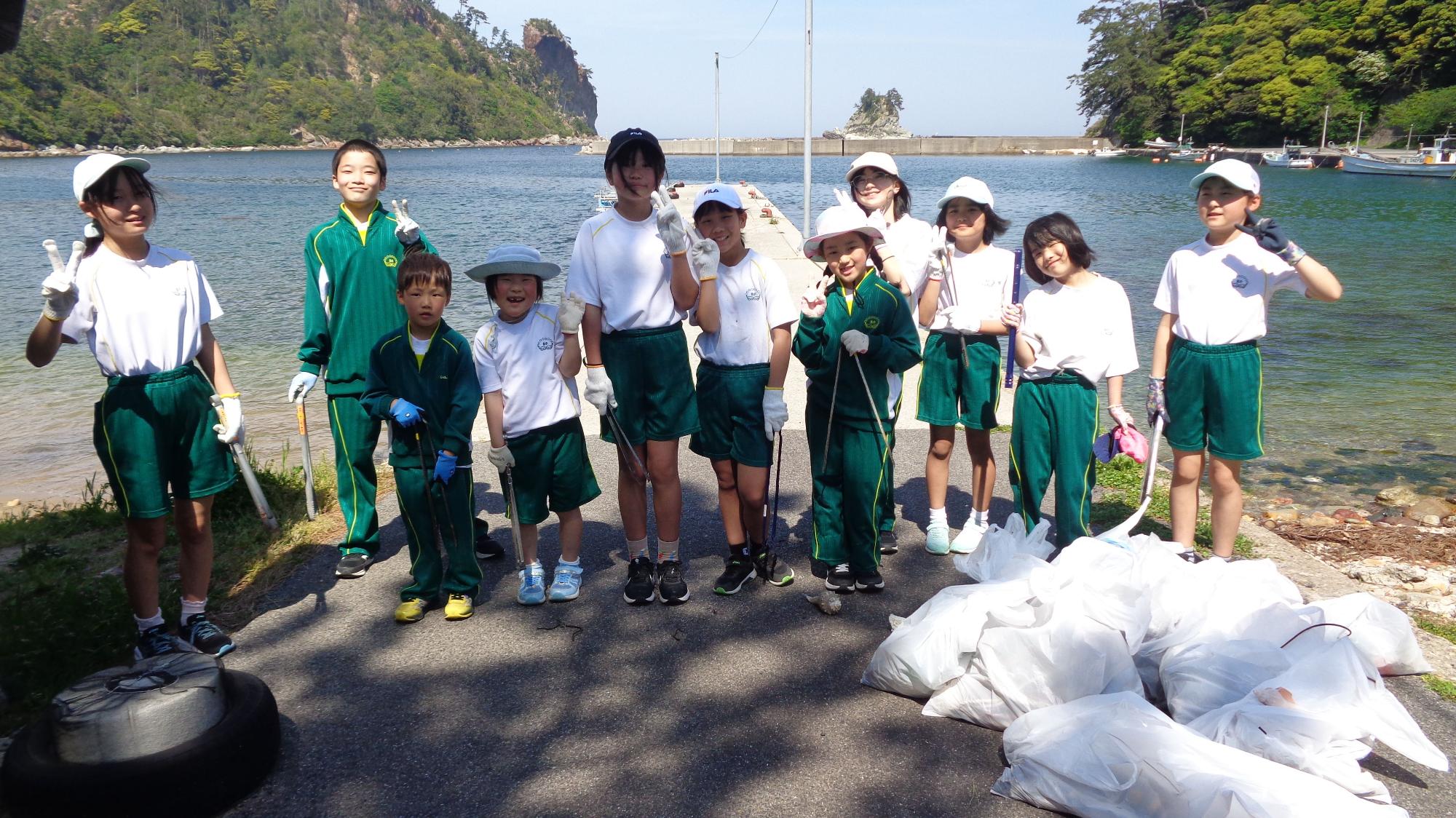海辺で体操着を着た児童たちがごみを集めてその前に集まりポーズをとっている写真