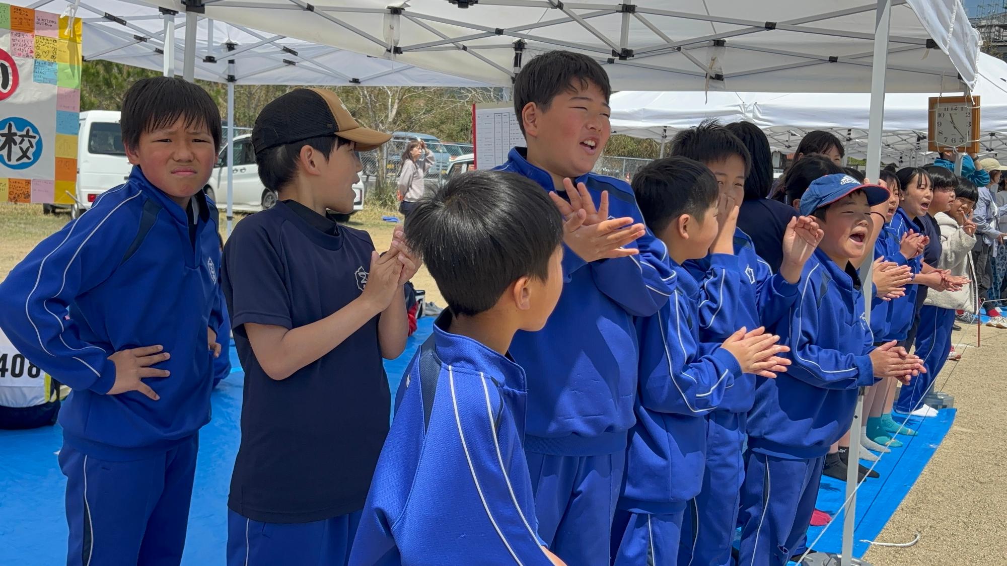 テントの下で青いジャージを着た児童たちが競技を応援しながら拍手をしている写真
