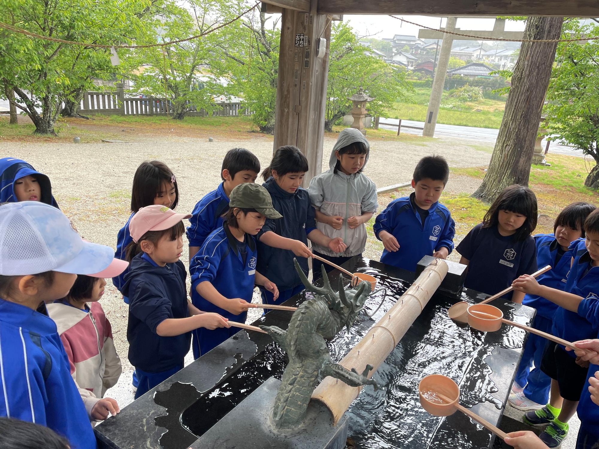 神社の手水舎で青いジャージを着た児童たちが柄杓で手を清めている写真