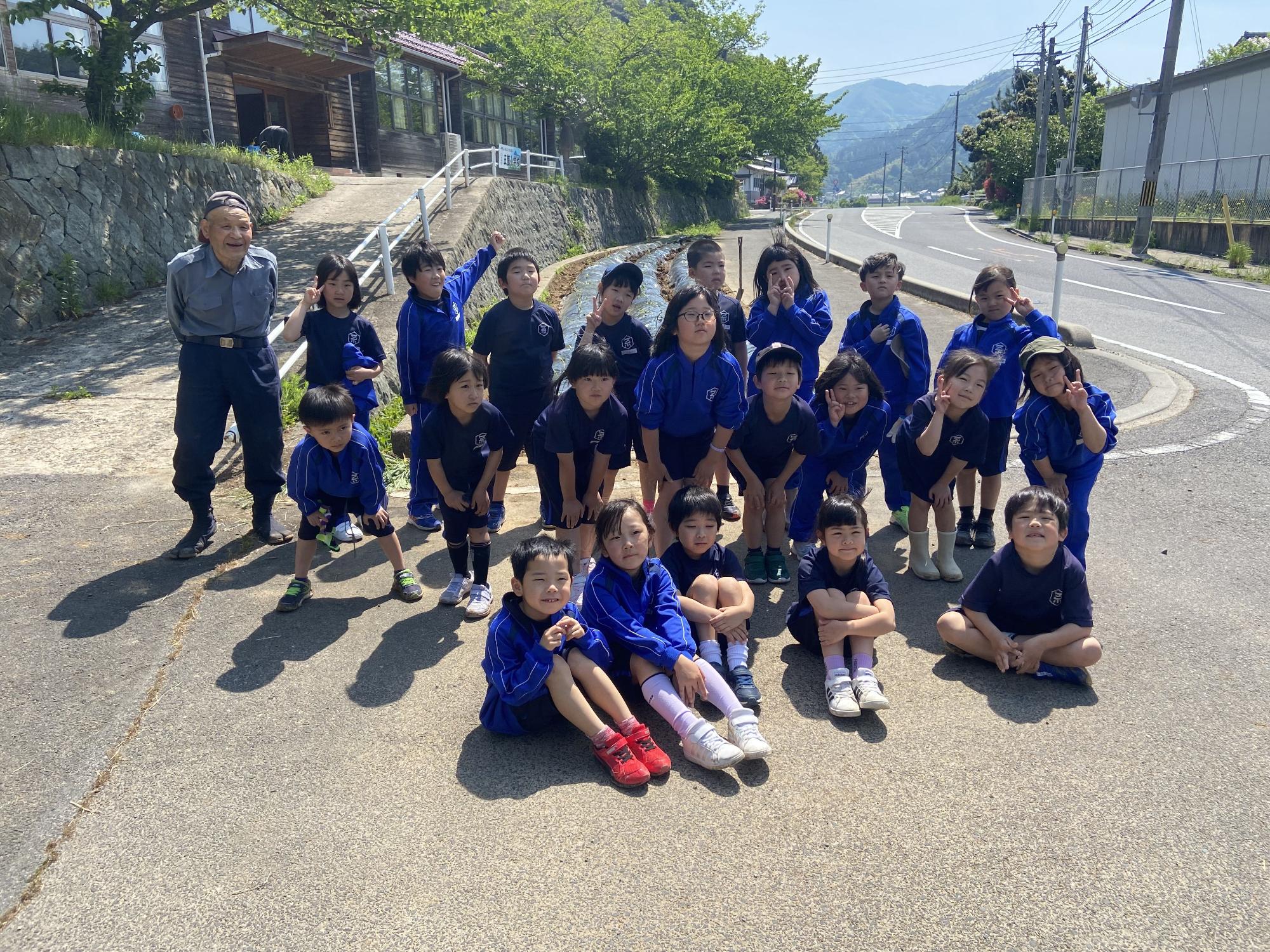 坂道のある校舎前で青い体操服を着た児童たちが並んで集合写真を撮っている写真