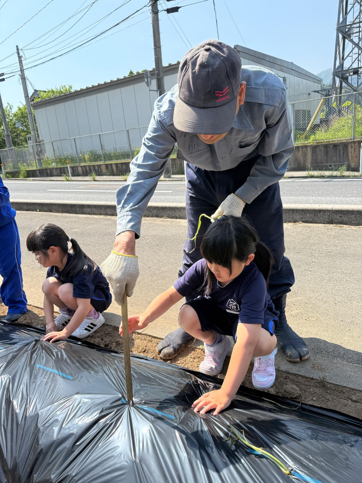 畑のそばで作業着を着た男性と児童が黒いマルチシートに穴を開けて苗を植えている写真