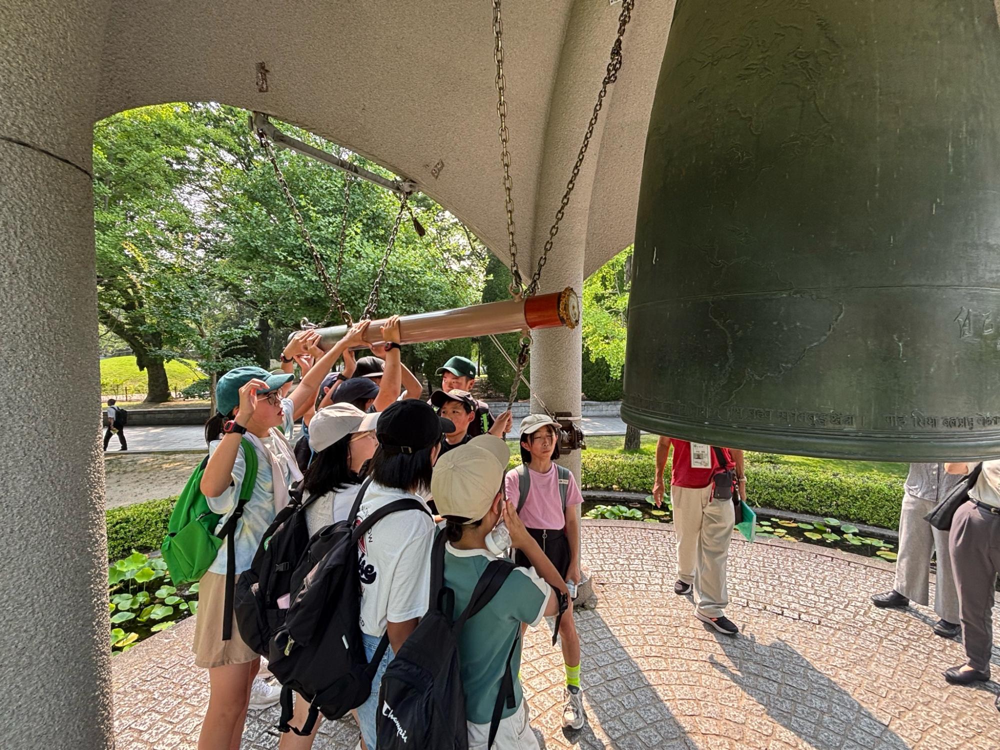 平和の鐘の下で帽子をかぶった児童たちが一緒に棒を持ち、鐘を鳴らしている写真
