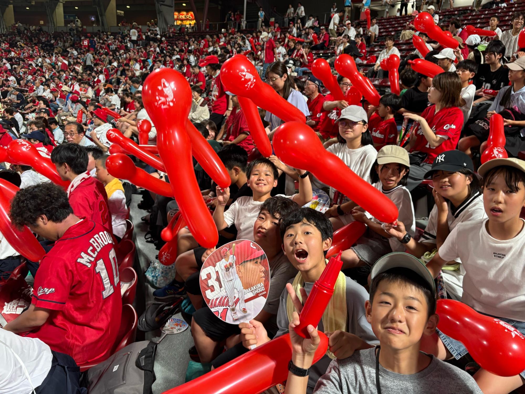 野球場のスタンドで赤い風船を持った児童たちが笑顔で応援している写真
