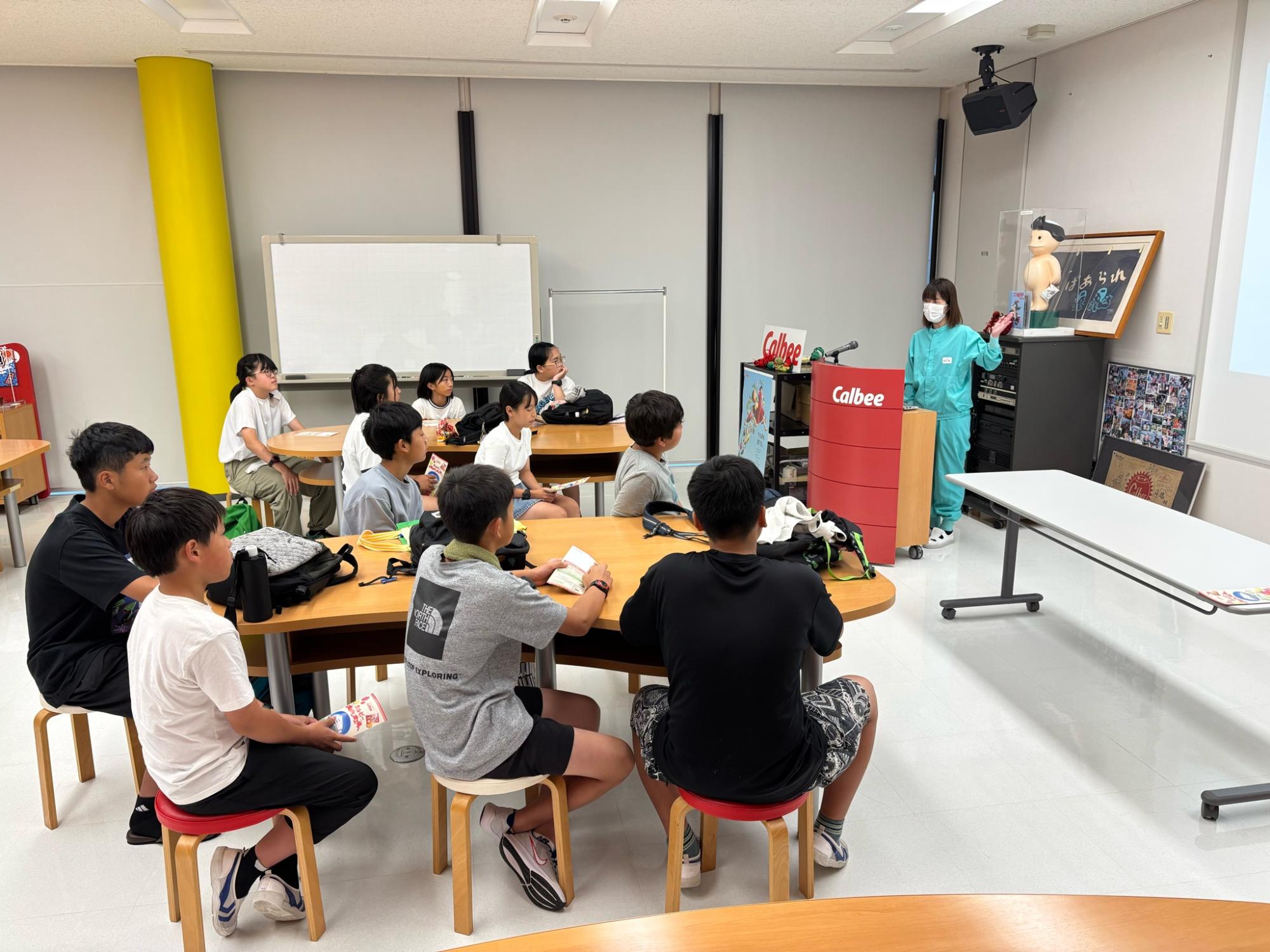 企業見学でマスクを着けた案内係の女性が児童たちに説明をしている写真