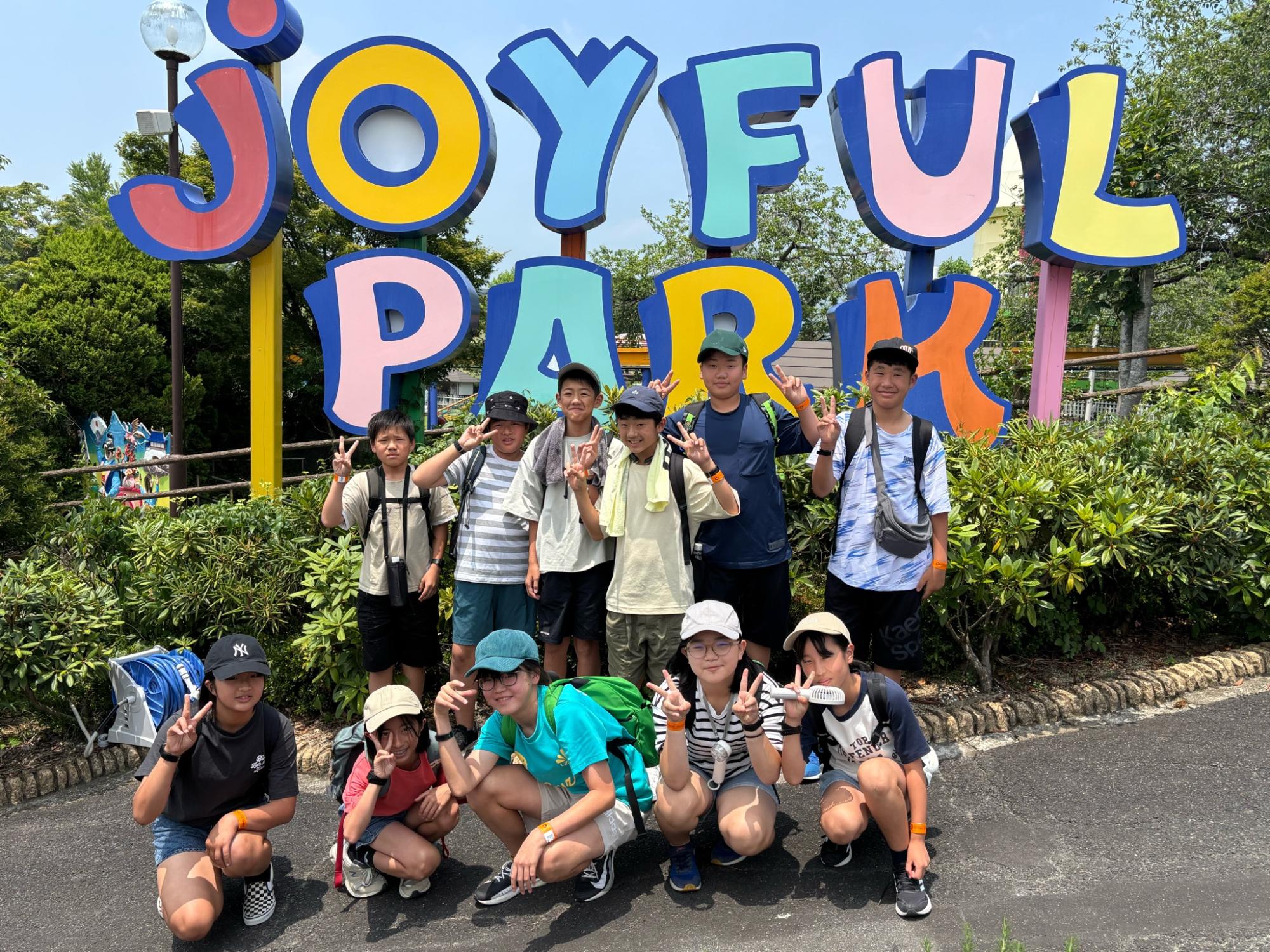 遊園地の入り口で「JOYFUL PARK」の看板を背に児童たちが笑顔でポーズをとっている集合写真