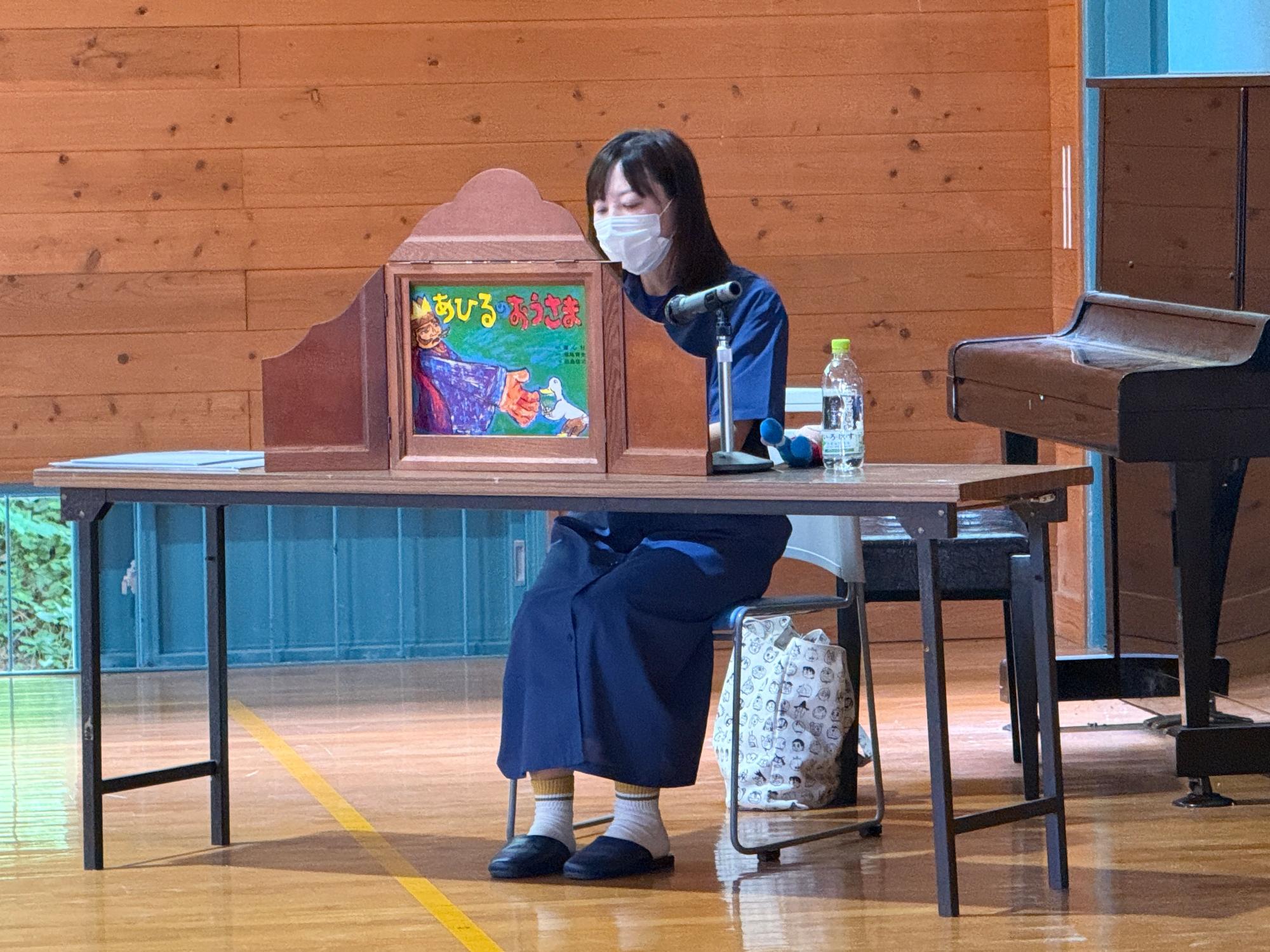 体育館で机の前に座ったマスクを着用した女性が紙芝居を読んでいる写真