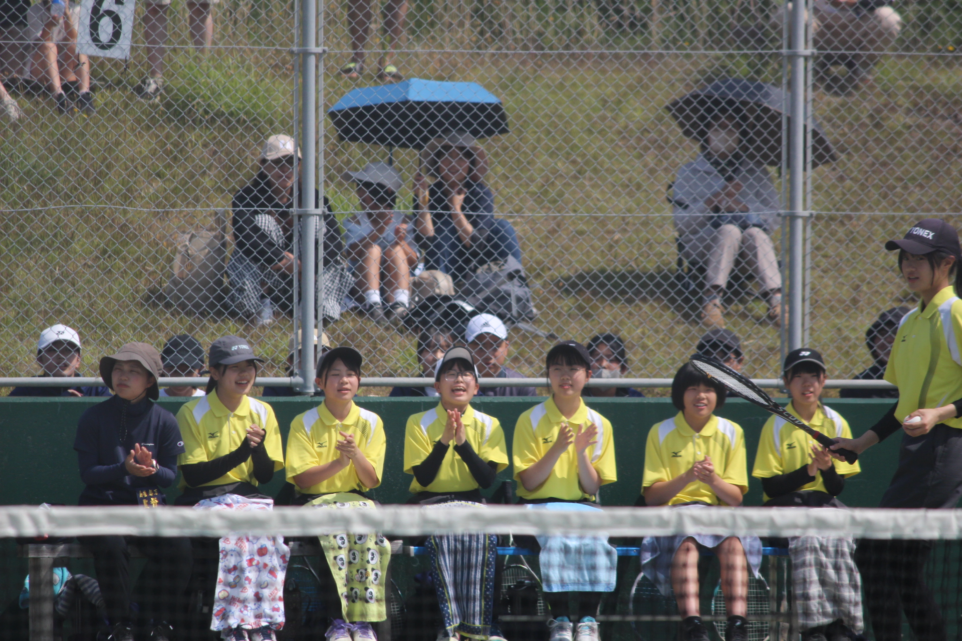観客席で黄色のユニフォームを着た生徒たちがコートの端でテニスの試合を応援している写真