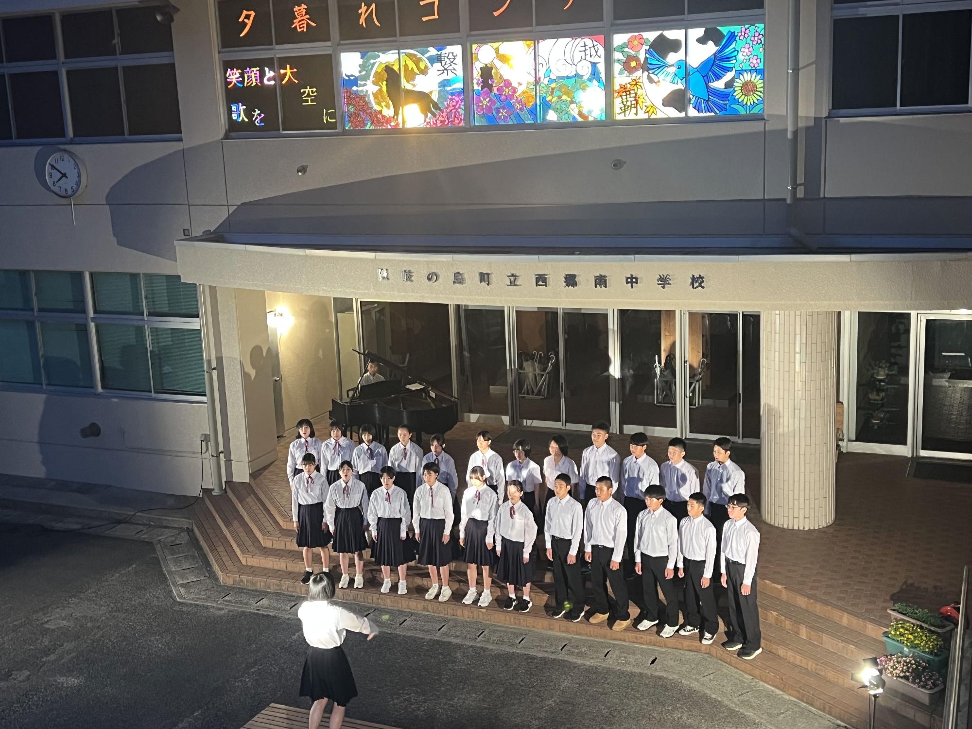 校舎前で生徒たちが指揮者に合わせて合唱をしている写真