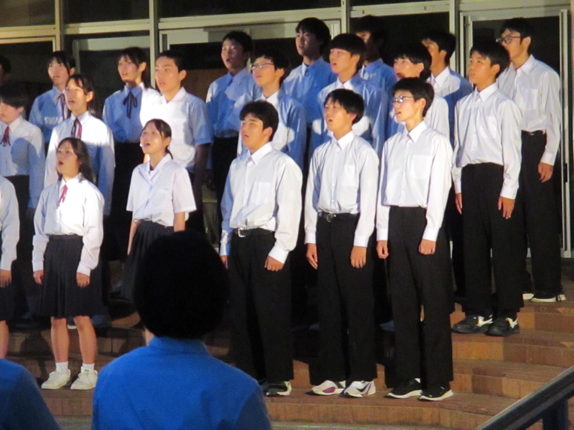 校舎前で生徒たちが階段に並んで合唱をしている写真