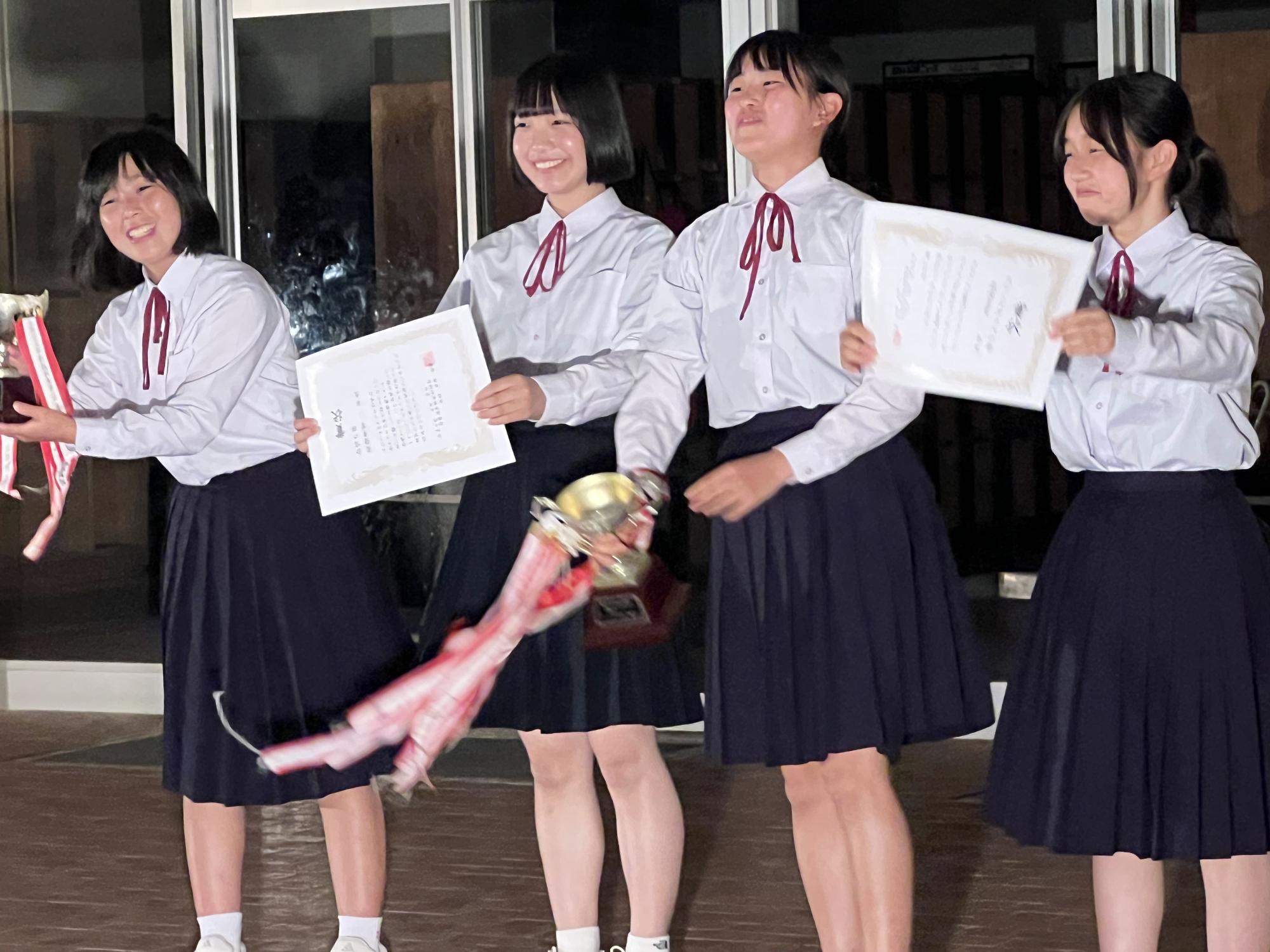校舎前で女子生徒たちが賞状やトロフィーを持ち笑顔を向けている写真