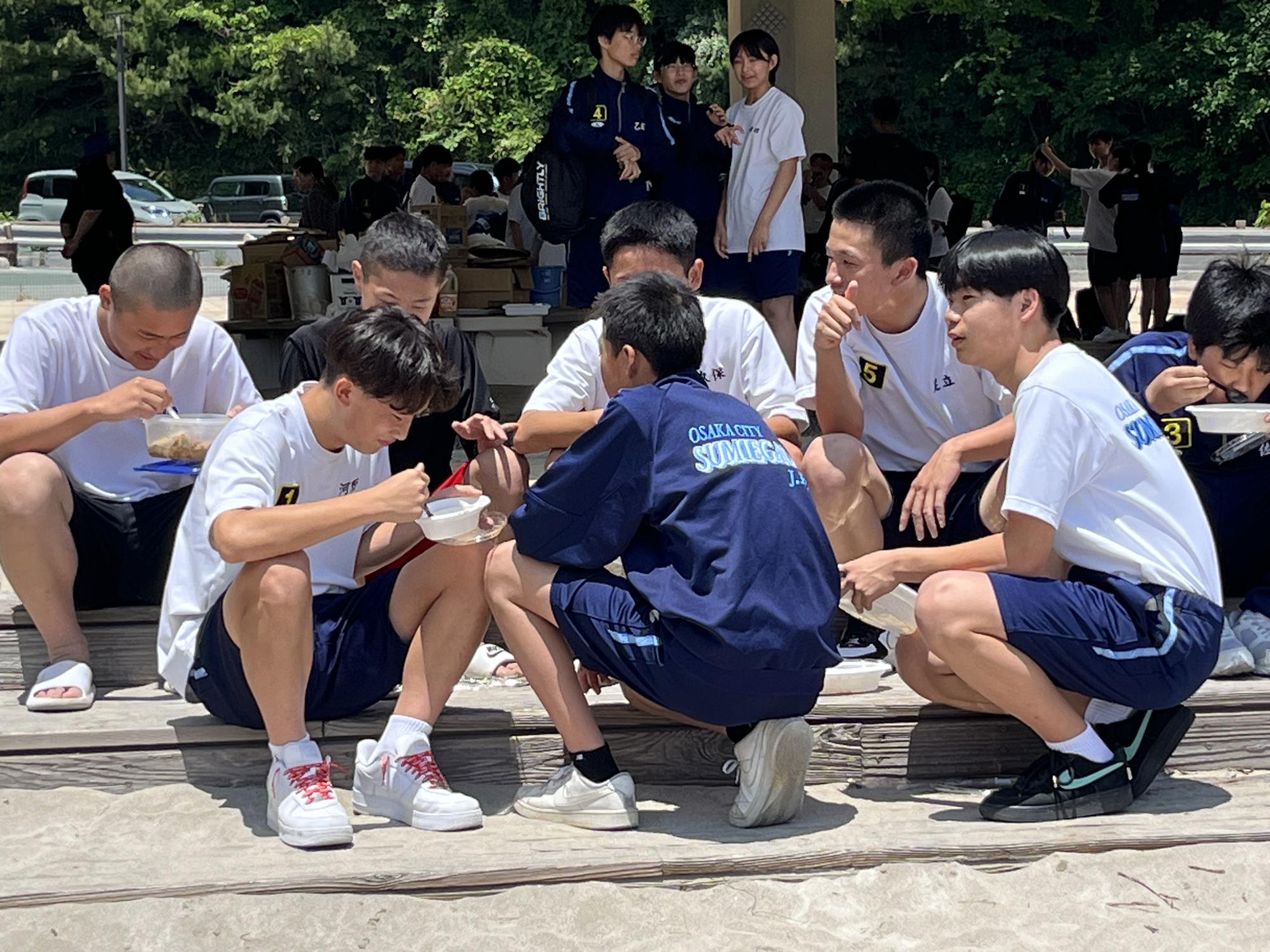 屋外で男子生徒たちがしゃがみ込みながら集まって昼食を食べながら談笑している写真