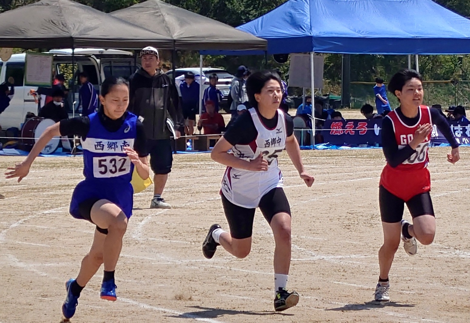 グラウンドで青色、白色、赤色のユニフォームを着た女子生徒たちが並走している写真