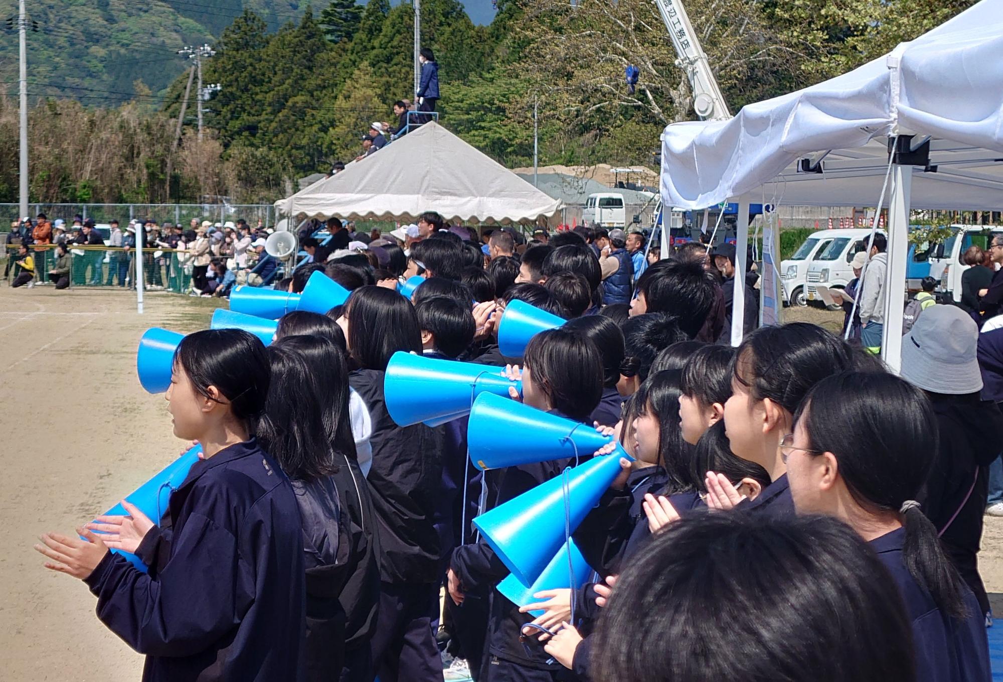 グラウンドで数十人の生徒たちが青いメガホンを持って声援を送っている写真