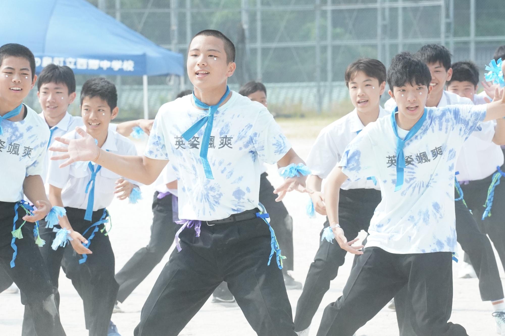 校庭で青い鉢巻を首に巻いた白Tシャツの生徒たちがダンスを披露している写真