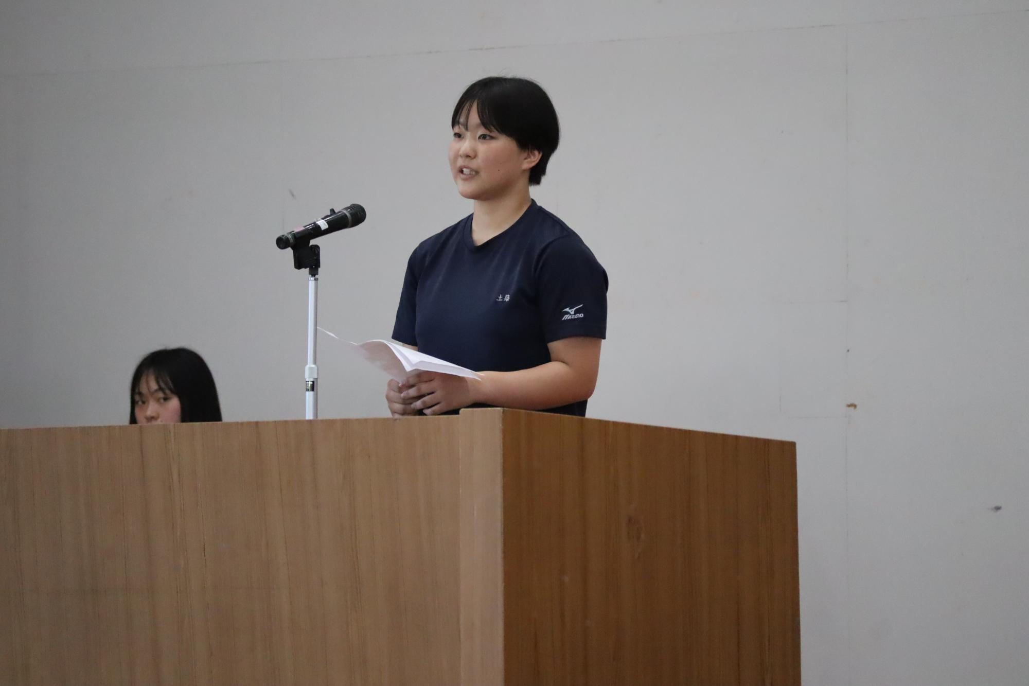 体育館で黒Tシャツを着た女子生徒が壇上からスピーチをしている写真