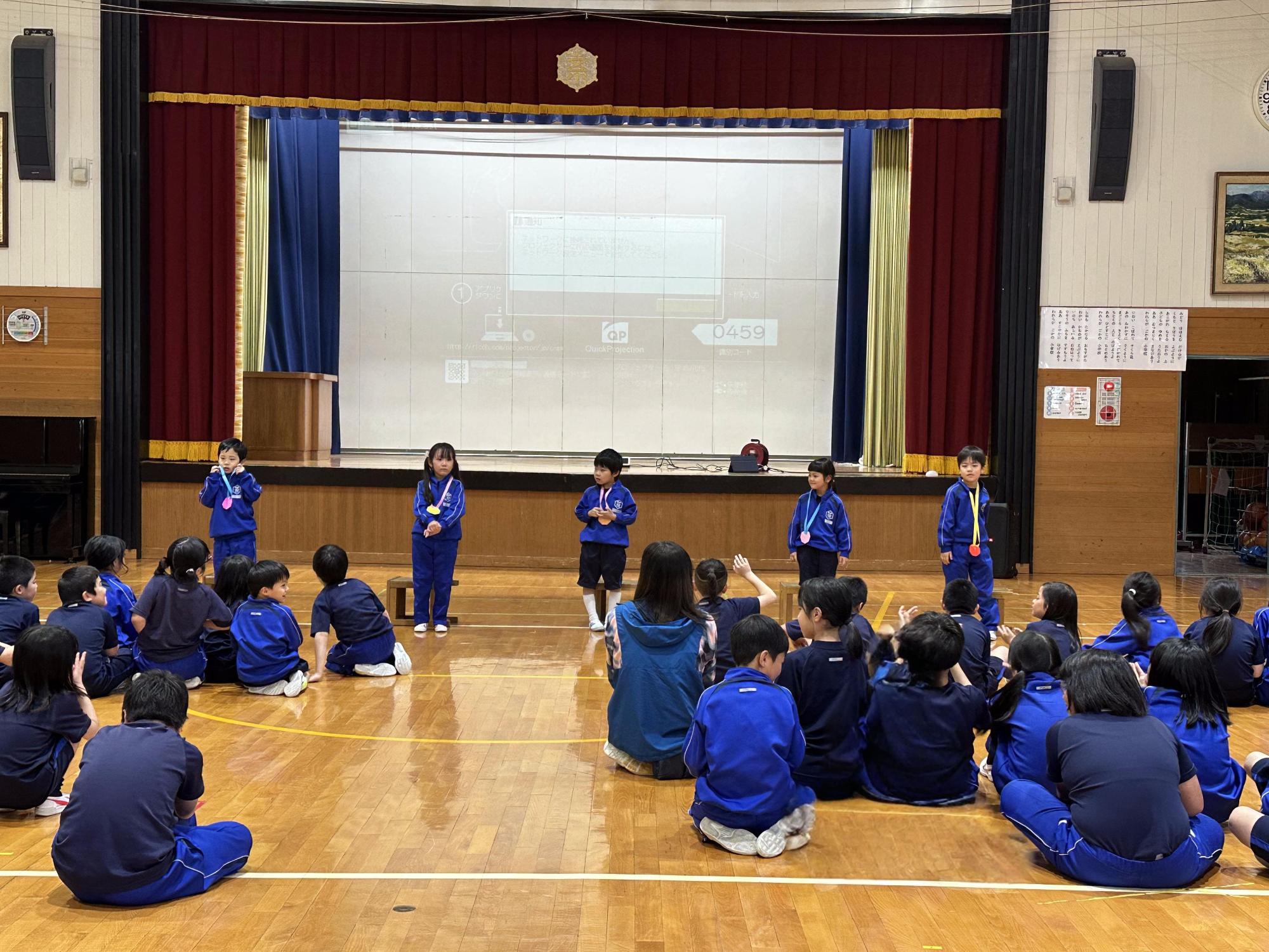 体育館で青いジャージを着た児童たちがメダルをかけて発表をしている写真