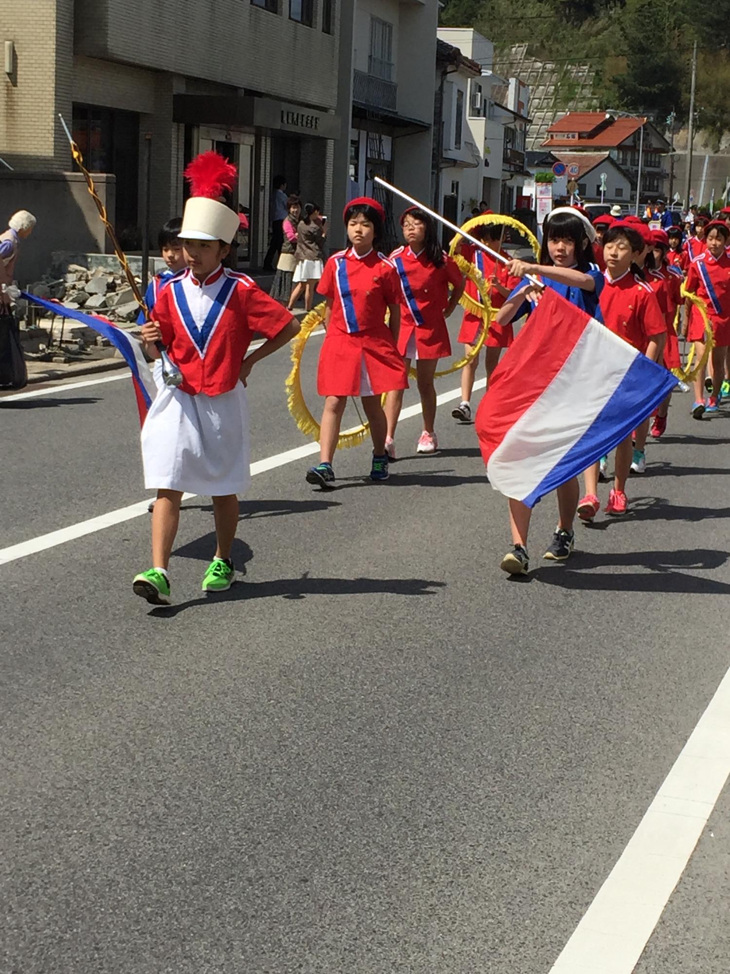 マーチングバンド風の衣装に身を包んだ児童が旗やバトンを手に持ち行進している写真