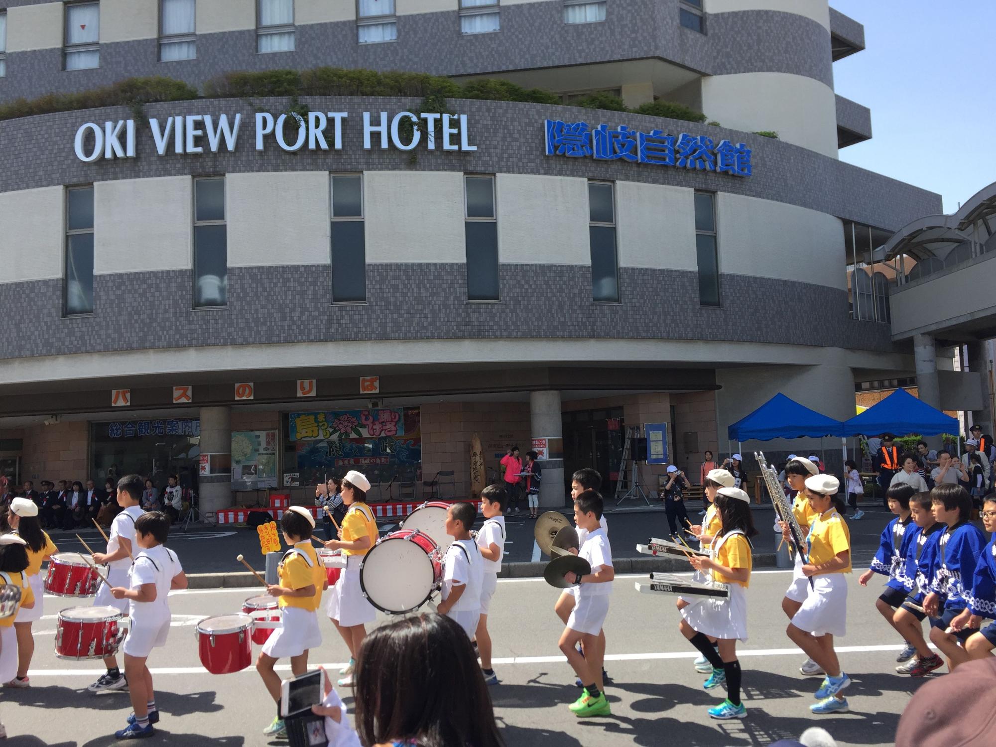 ホテル前の道路を、楽器を持った児童たちが鼓笛隊として行進している写真