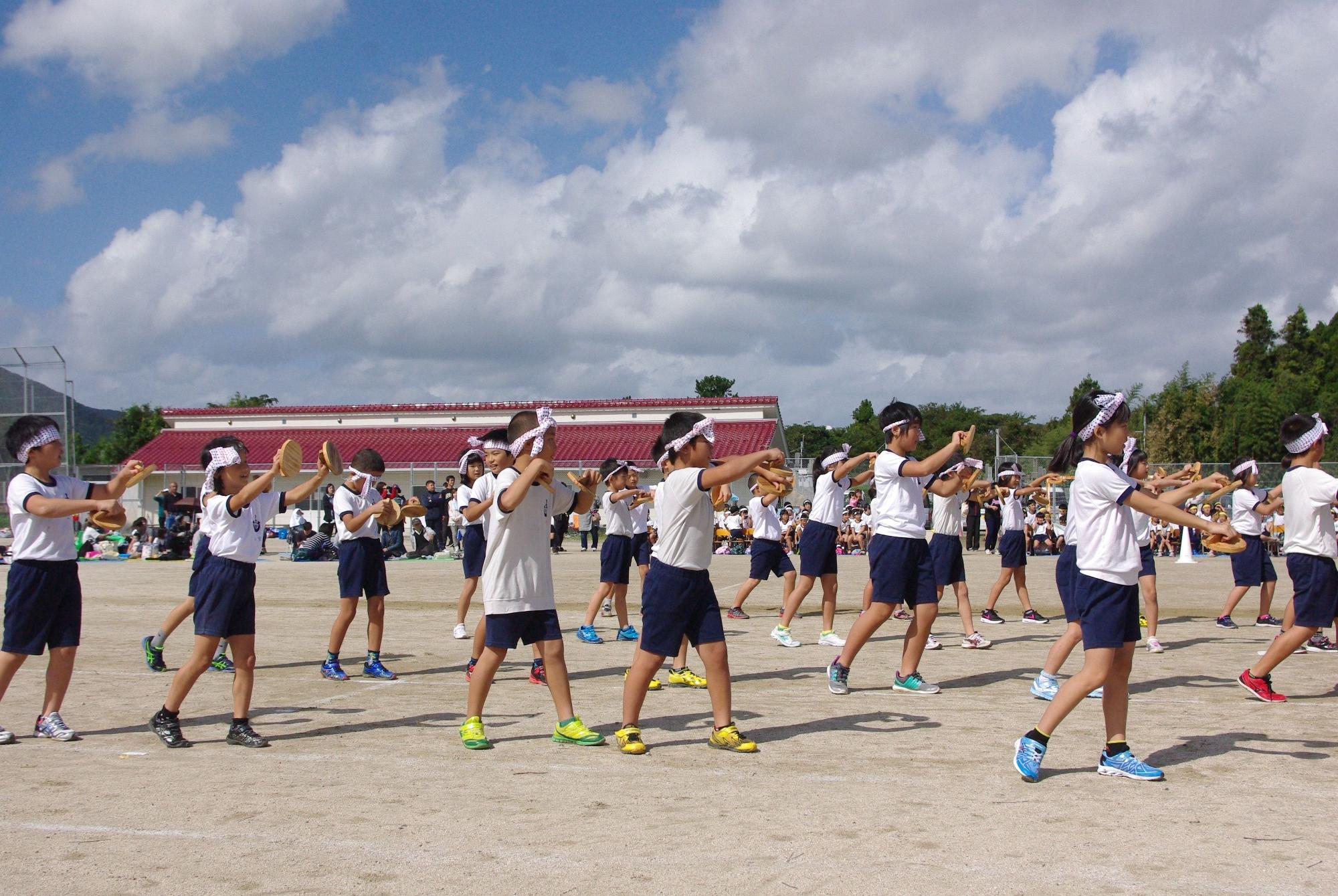 校庭でハチマキに体操服姿の児童が木製の小道具を手に持って踊りを踊っている写真
