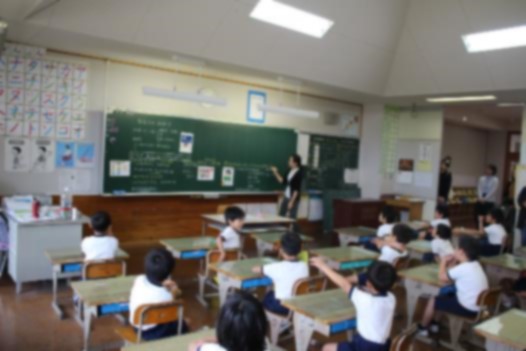 児童たちが自分の席で黒板に字を書いてる先生の授業を聞いている写真