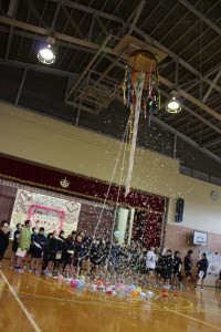体育館で児童達が天井からくす玉がぶら下がっておりそこから割れた飾りを見上げている写真