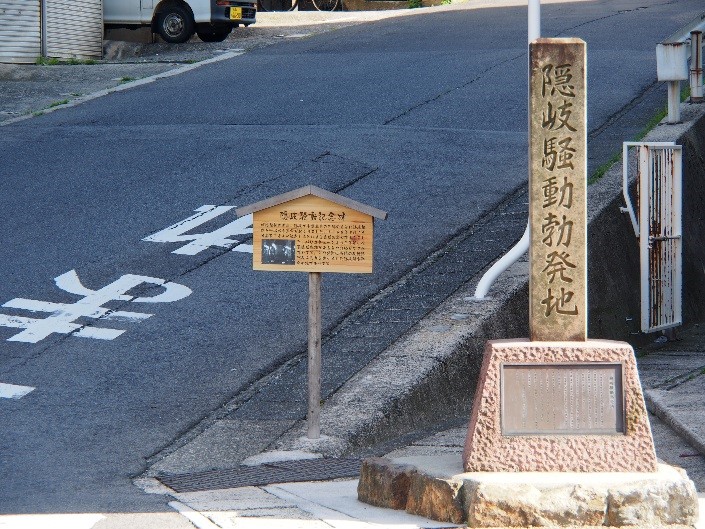 「隠岐騒動勃発地」と彫られた石碑と木製看板が道路の脇に備えられている写真