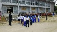 校舎前で運動着を着た児童たちが整列して話を聞いている写真
