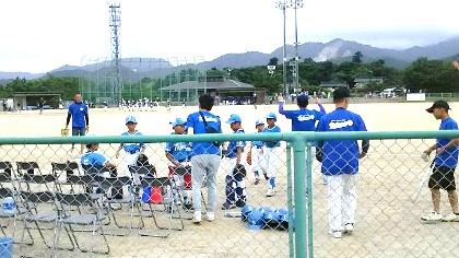 フェンス越しに学童ソフトボールチームが監督のもとに集合している写真