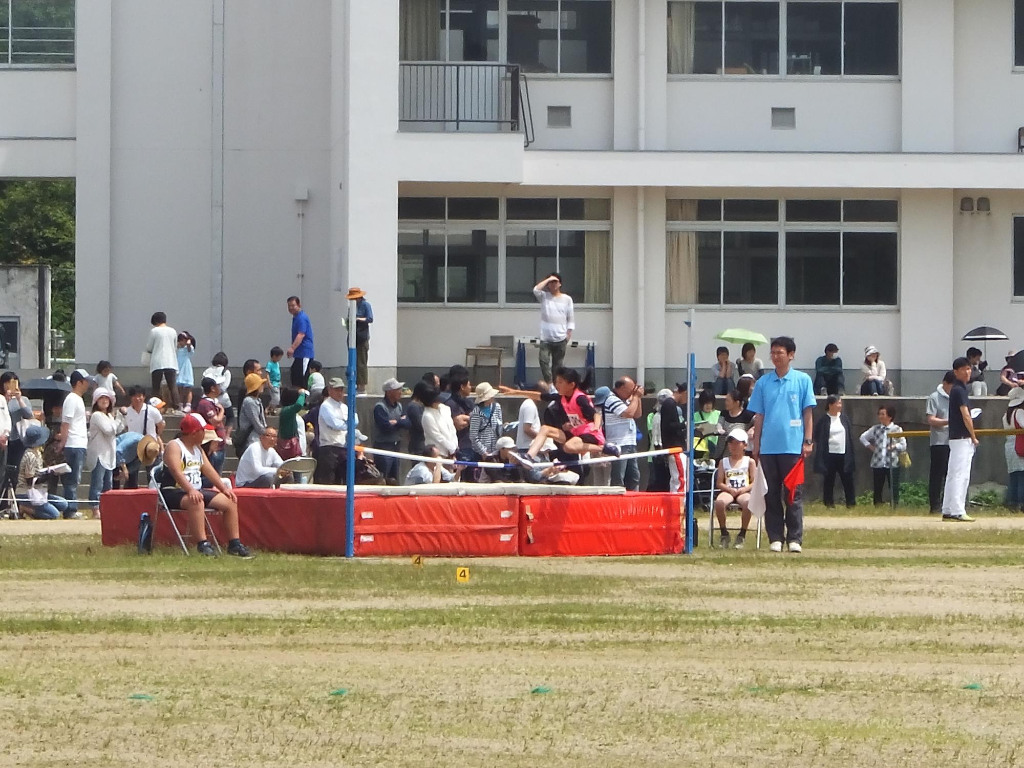 グラウンドで1人のユニフォーム姿の児童が走り高跳びを行っている様子を沢山の人が見ている写真