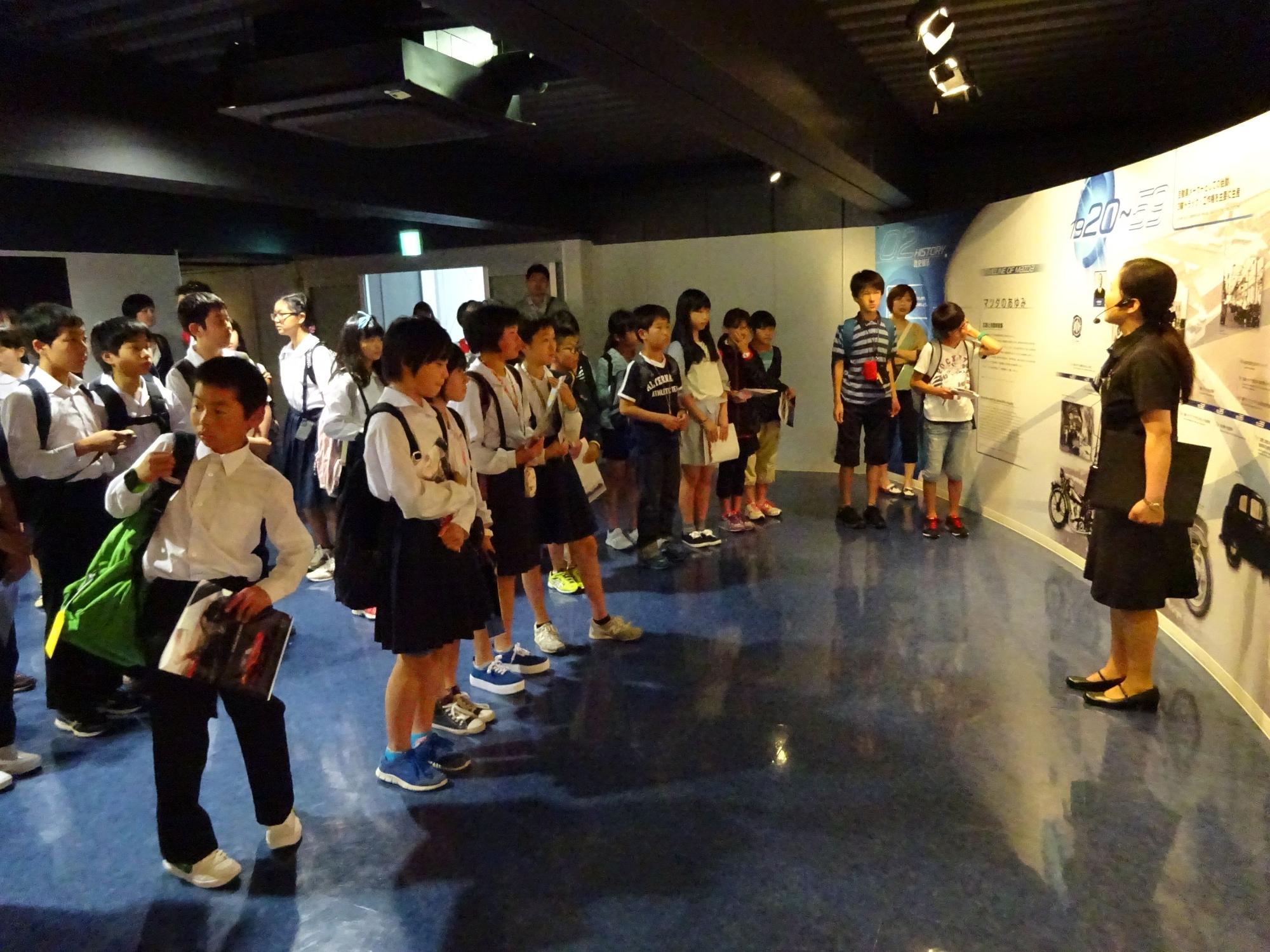 ヘッドマイクを付けた女性の説明を複数の児童が聞いている写真