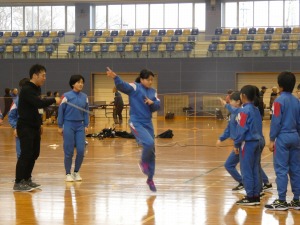 体育館で男性や児童に見守られながら中央の1人の児童が跳躍している写真