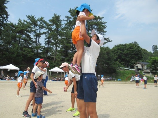 体操着姿の背の低い児童が上級生に抱き上げられている写真