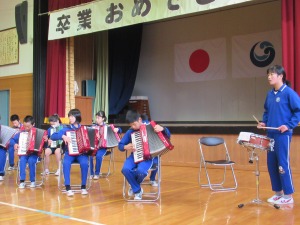 体育館のステージ前で複数の児童が椅子に座り、楽器を演奏している写真