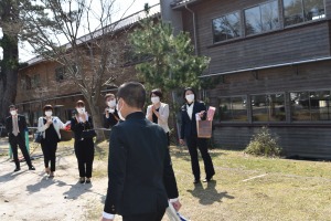 校舎の前で児童が歩き、複数の男女が並んで拍手で見送っている写真