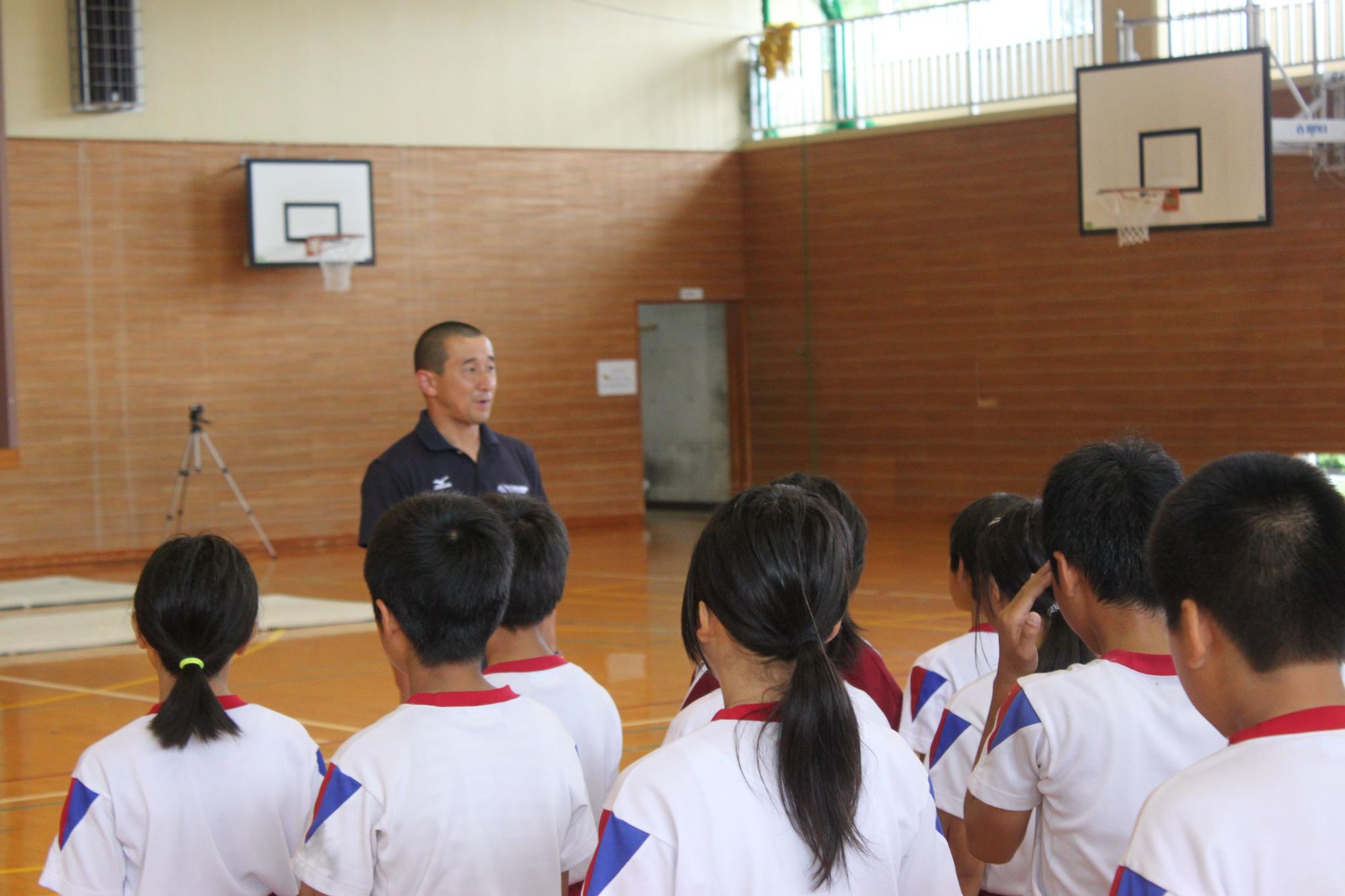 体育館で児童たちが男性の話を聞いている写真