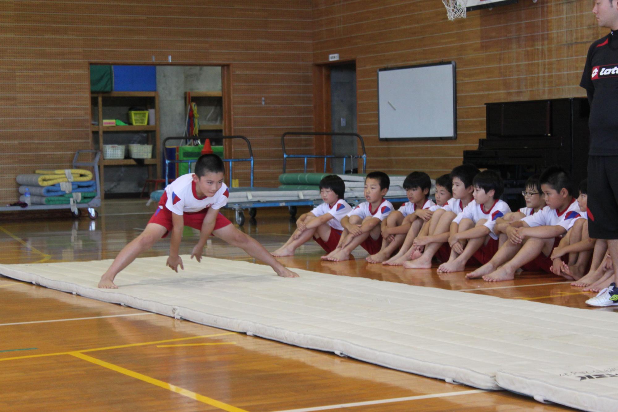 体育館に敷かれたマットの上でポーズを決めている男子児童を複数の児童が見守っている写真