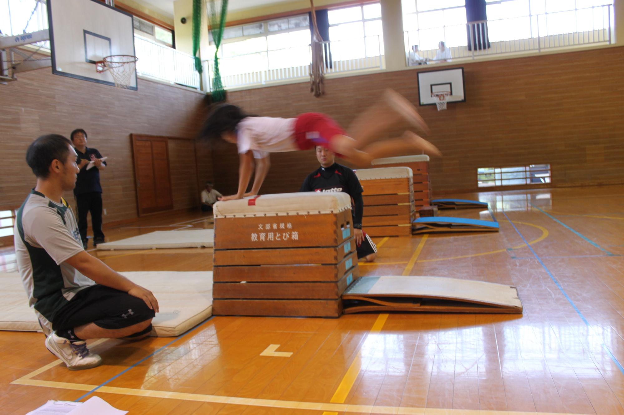 体育館で跳び箱を行っている児童を男性が見守っている写真