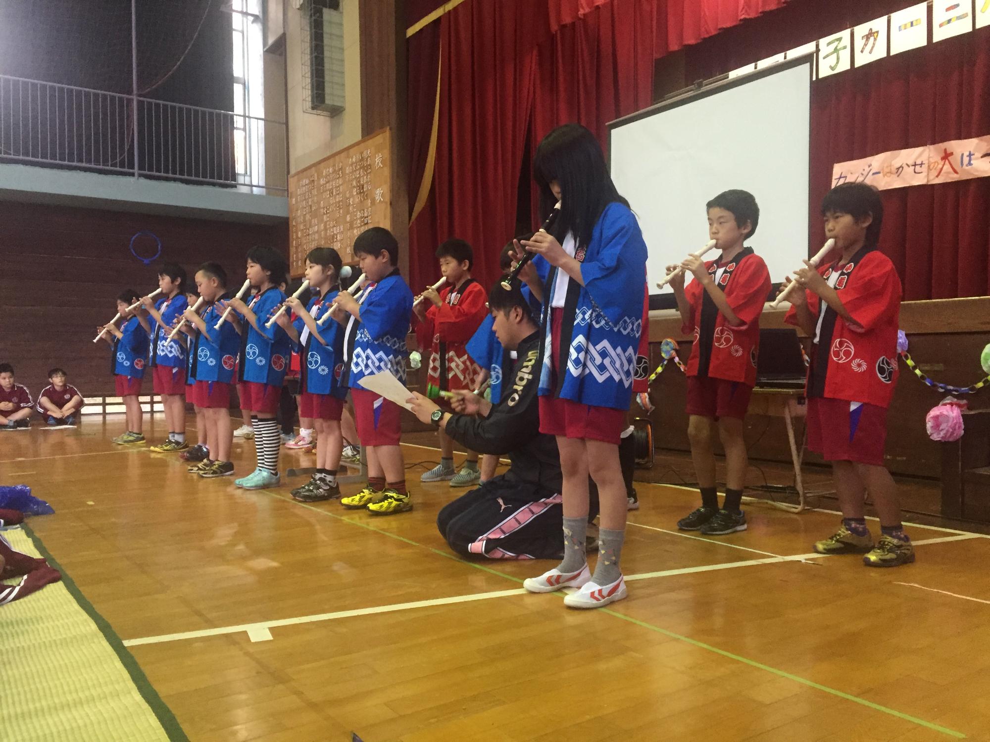 体育館のステージ手前で法被姿の複数の児童がリコーダーを演奏している写真