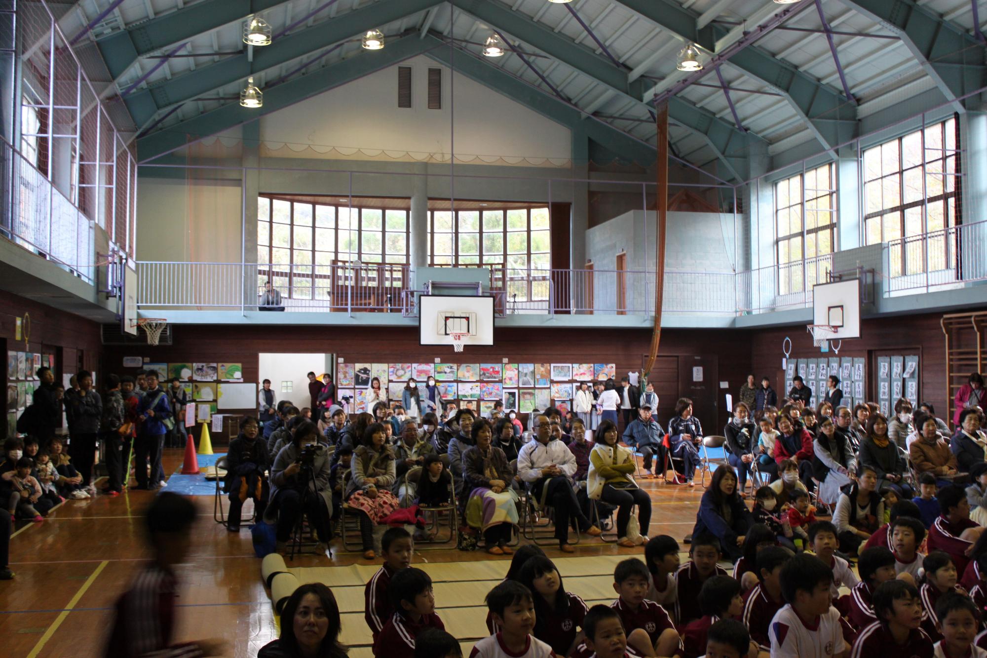 体育館で児童が前方に座り、後方には多くの人々が椅子に腰掛けている写真