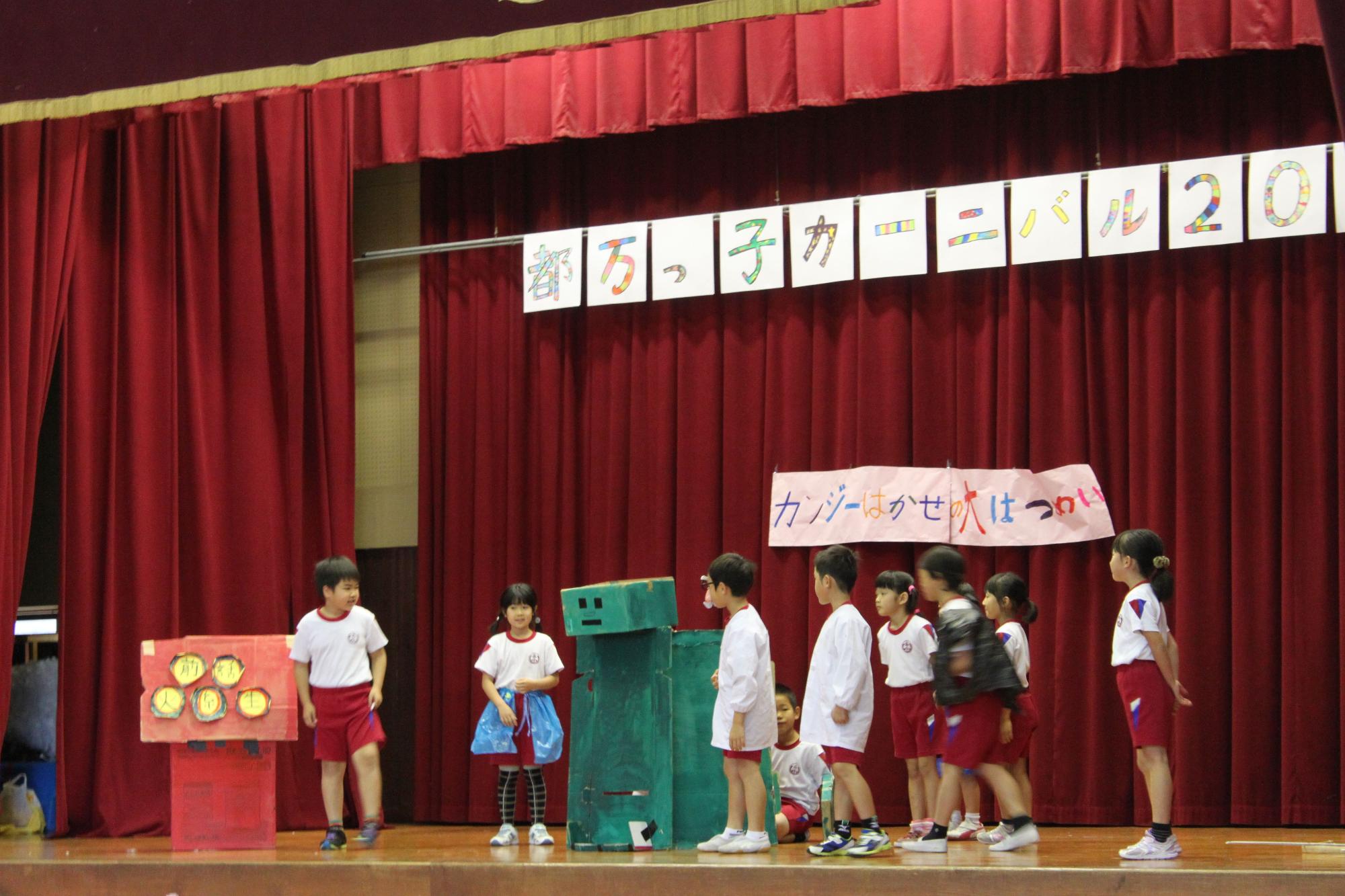 ステージ上で複数の児童たちが小道具を使いながら劇を演じている写真