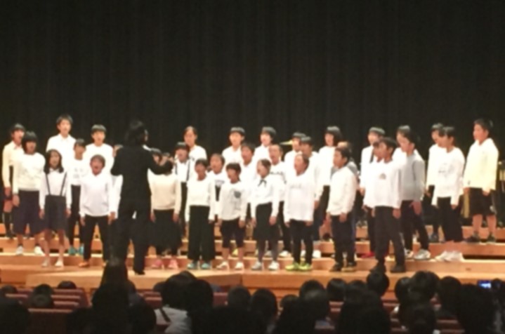 舞台で児童が指揮者に合わせて合唱している写真