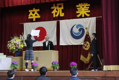 ステージの壇上で、卒業生が一人ずつ校長先生から卒業証書を受け取っている写真