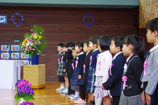 体育館で新入生の児童たちが左胸にバッチをつけ並んでいる写真