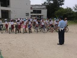外でヘルメットを被り自転車の横に立ち、警察署の方の話を聞いている児童たちの写真