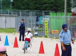 ヘルメットをかぶり自転車に乗り、置かれた三角コーンに沿って走っている児童を警察が見守っている写真